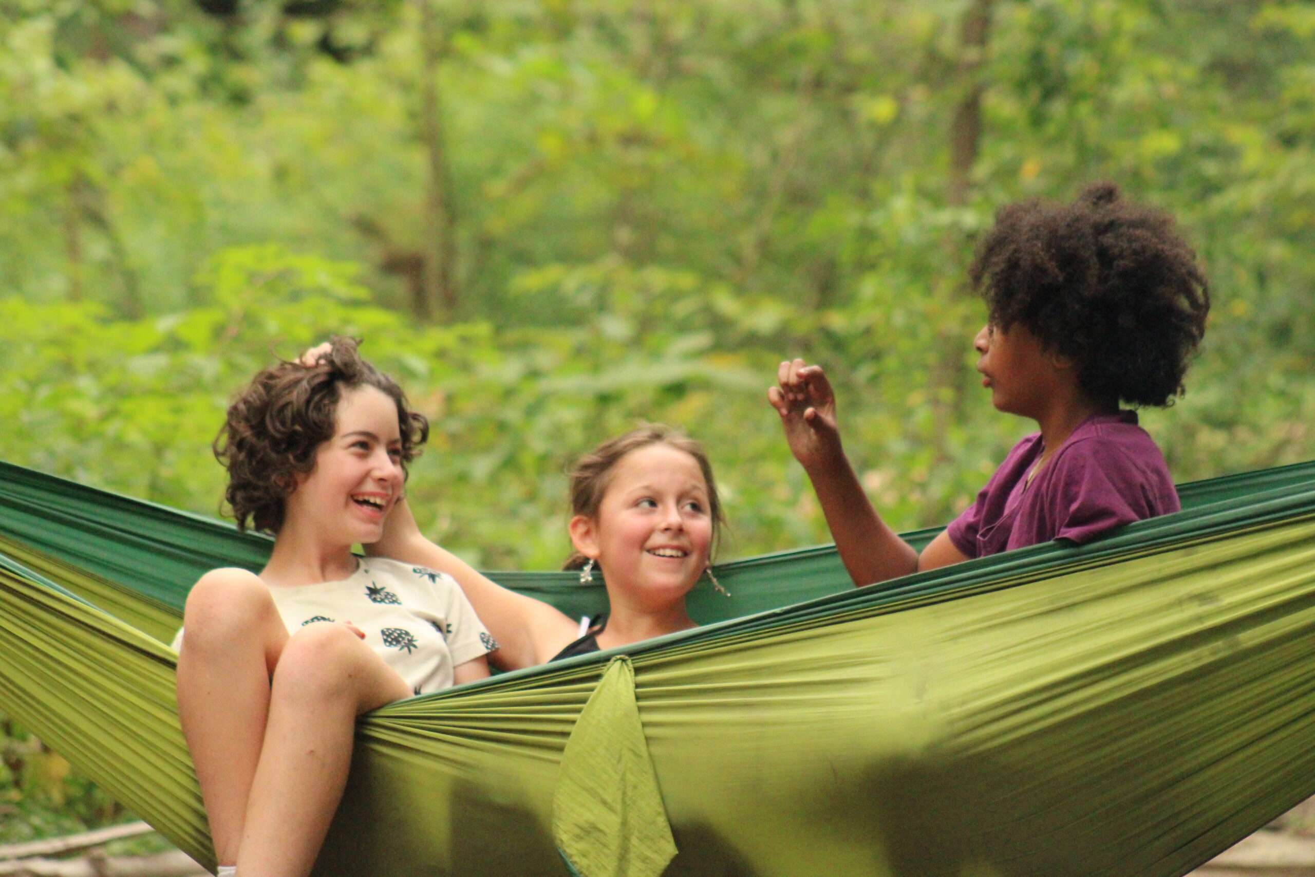 three young girls sitting in a hammock all smiling and talking 