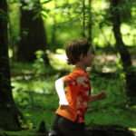 A young boy in an orange shirt running through the woods.