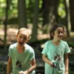 Two children, both smiling and both in light blue shirts, standing in the forest.