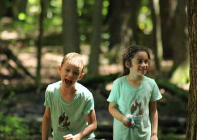 Two children, both smiling and both in light blue shirts, standing in the forest.