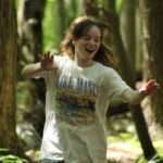 A smiling teenager with her hands up running through a leafy forest.