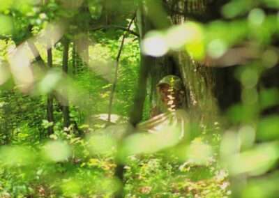 A leafy foreground in a forest with a smiling person leaning against a tree.