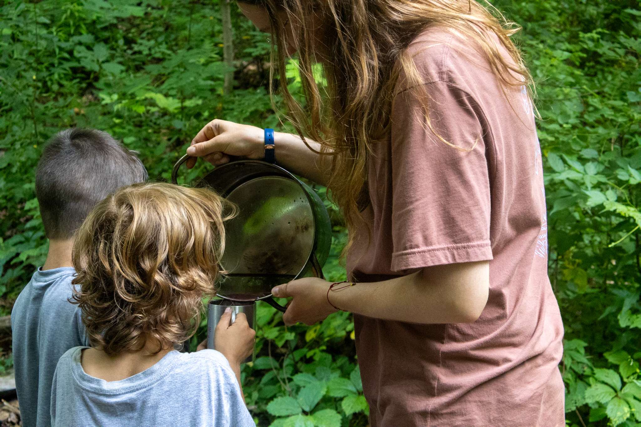 Instructor pouring beverage from pot into cups held by two campers.