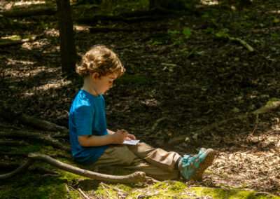 Young camper sitting on the forest floor, drawing in a small journal.