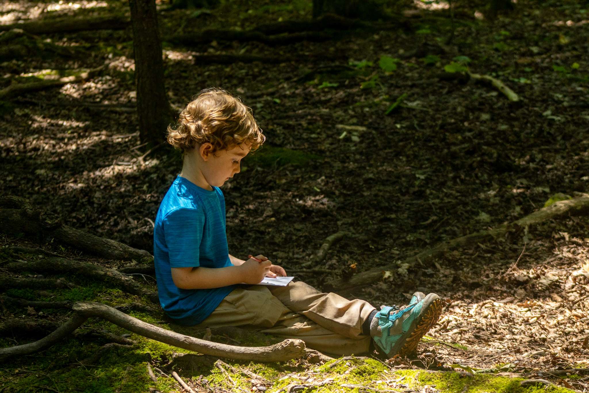 Young camper sitting on the forest floor, drawing in a small journal.