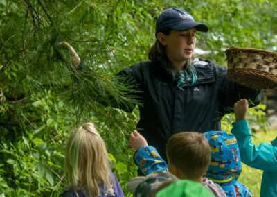 An instructor holding a pine tree branch and a woven basket while speaking to younger campers.