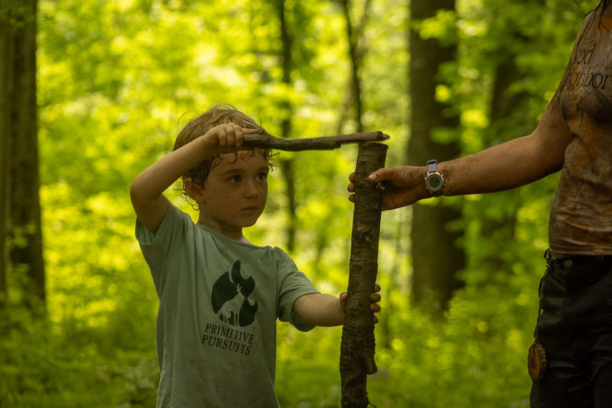 A child using a small stick to hammer a larger stick into the ground while an instructor holds the larger stick in place.