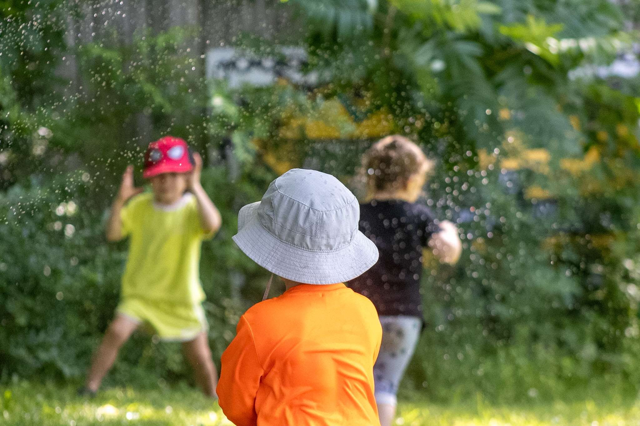IMG_9566 A young boy watching two other younger campers run under a sprinkler outdoors.