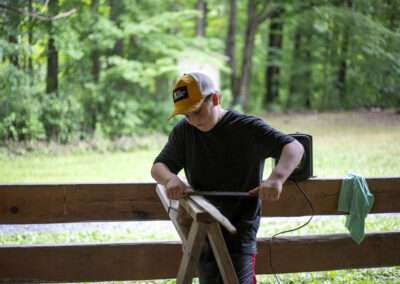 An older camper holding a wooden board on a flat wooden surface, tying rope around the board.