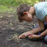 A boy kneeling and lighting a small pile of twigs on fire with a match.