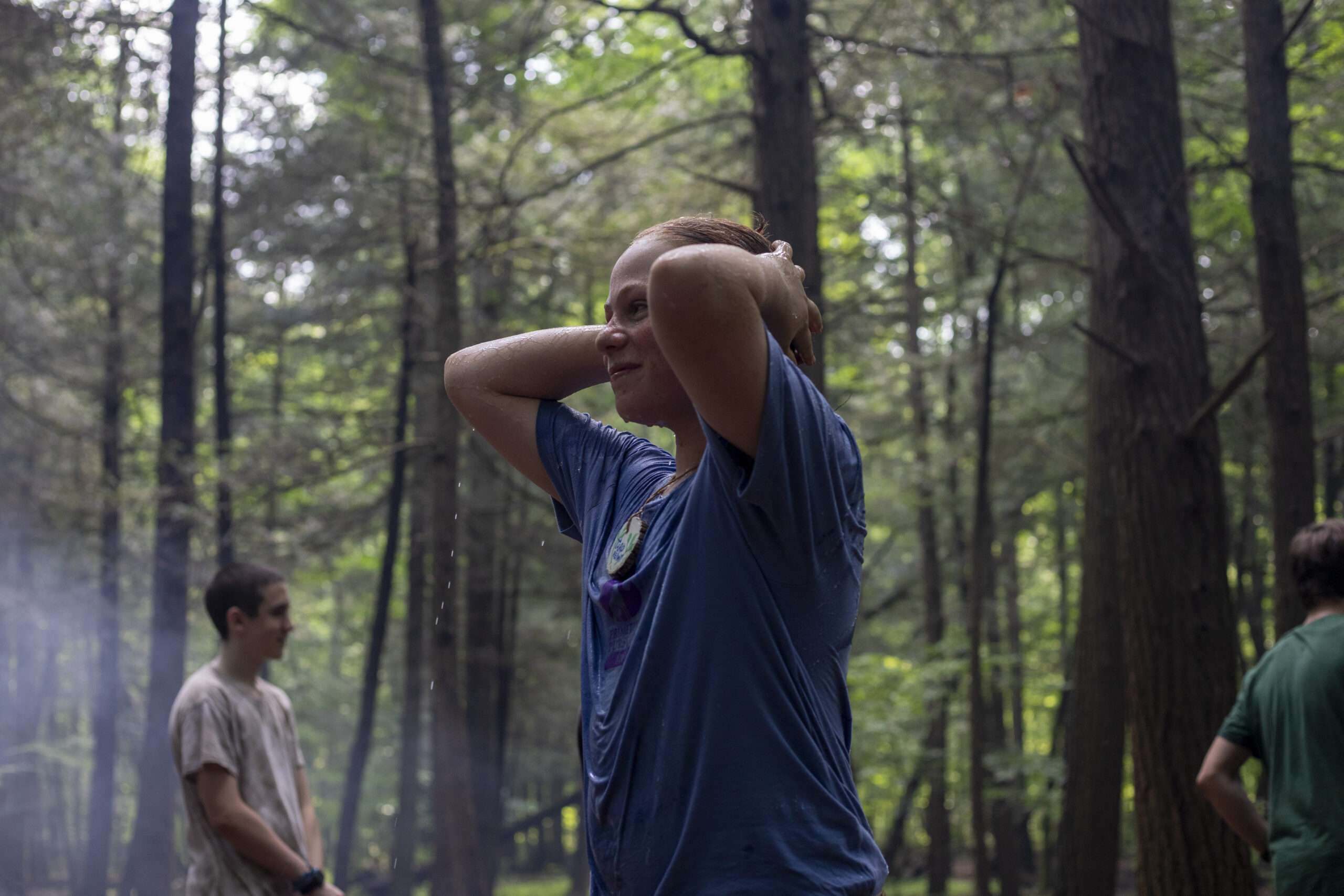 An older camper standing with hands on head in the woods, two other older campers are visible in the background.