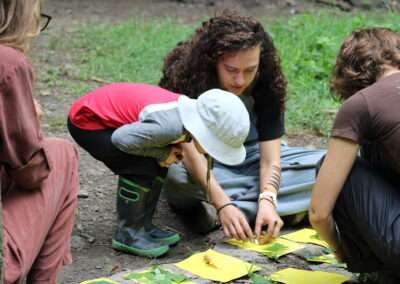 A younger camper observing two instructors as they lay out leaves on different pieces of paper.