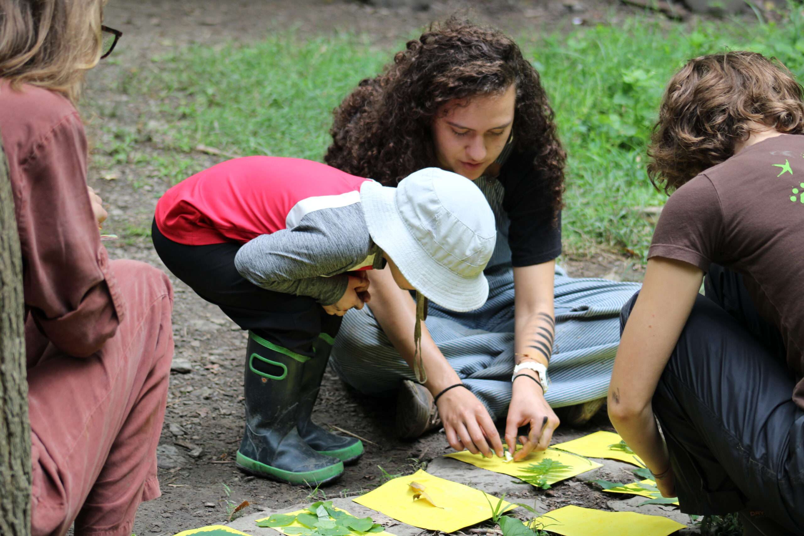 A younger camper observing two instructors as they lay out leaves on different pieces of paper.